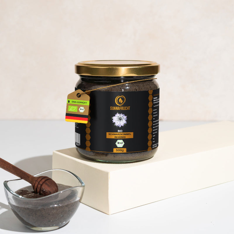 Glas Bio Schwarzkümmel in Honig von Sunnafrucht®, 500 g, auf cremefarbenem Podest mit Schälchen der dunklen Honig-Schwarzkümmel-Mischung und Holzlöffel