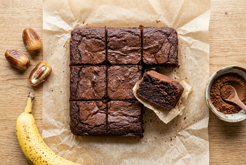 Brownies mit Datteln und Bananen in neun Stücke geschnitten auf Backpapier, daneben Rutab Datteln, eine reife Banane und eine Schale mit rohem Kakao