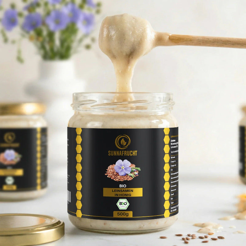 Geöffnetes Glas Bio Leinsamen in Honig von Sunnafrucht® mit Holzlöffel, der die cremige Mischung hebt, mit Leinblüten und Leinsamen