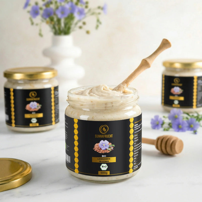Geöffnetes Glas Bio Leinsamen in Honig von Sunnafrucht® mit cremig gewirbelter Mischung und Holzlöffel, daneben Leinblüten und Honiglöffel