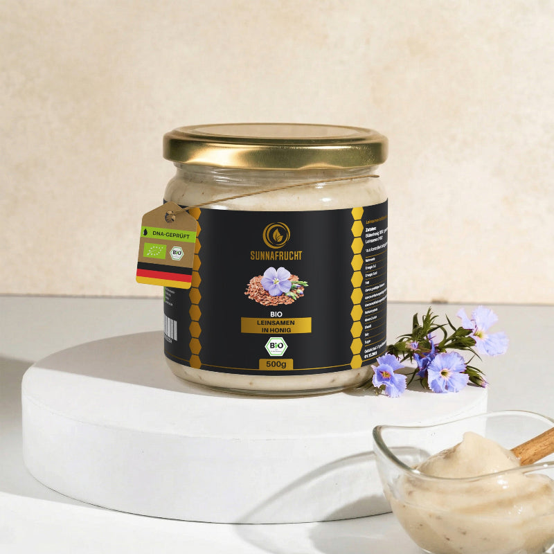 Glas Bio Leinsamen in Honig von Sunnafrucht®, 500 g, auf weißem Podest mit blauen Leinblüten und Schälchen der cremigen Honig-Leinsamen-Mischung