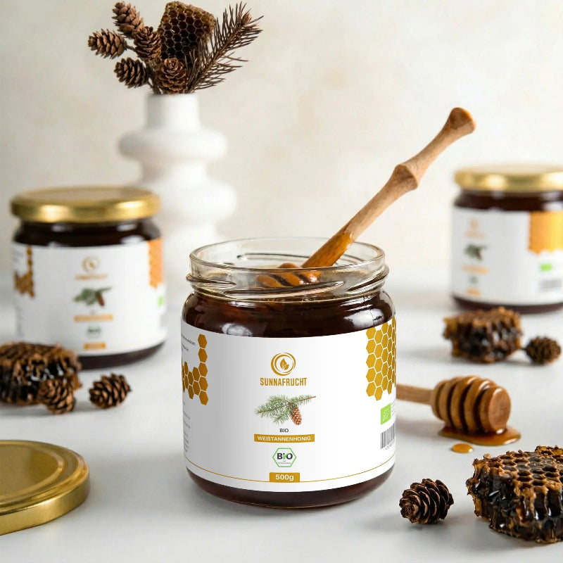 Geöffnetes Glas Bio Weißtannenhonig von Sunnafrucht® mit Holzlöffel, umgeben von Tannenzapfen, Wabenstücken und Honiglöffel