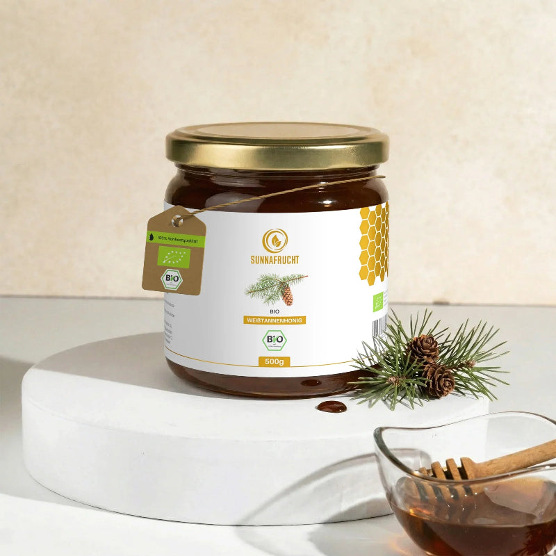 Glas Bio Weißtannenhonig von Sunnafrucht®, 500 g, auf weißem Podest mit Tannenzweig, Zapfen und Schälchen dunklen Honigs mit Honiglöffel