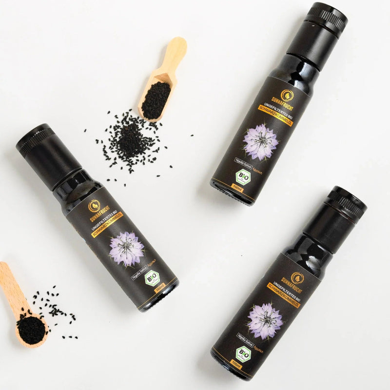 Drei Flaschen Bio Schwarzkümmelöl und Holzlöffel mit Schwarzkümmelsamen auf weißem Hintergrund